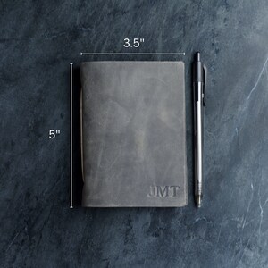 May include: A gray leather journal, 5 inches tall and 3.5 inches wide, with the initials "JMT" embossed on the cover. A black pen rests beside the journal. The journal has a worn, textured appearance.