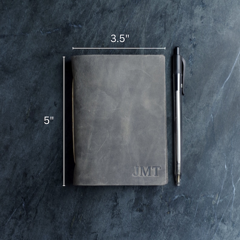 May include: A gray leather journal, 5 inches tall and 3.5 inches wide, with the initials "JMT" embossed on the cover. A black pen rests beside the journal. The journal has a worn, textured appearance.