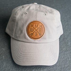 May include: A light gray baseball cap featuring a circular brown leather patch centered on the front. The patch displays an embossed compass design with arrows pointing outward and the letters N, E, S, and W positioned between the arrow shafts.