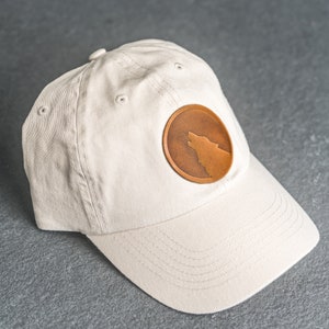 May include: A beige baseball cap with a brown leather patch featuring a wolf silhouette.