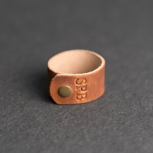 Op de afbeelding: Een beige leren manchetarmband met een gladde textuur en een ronde, messingkleurige metalen drukknoopsluiting. Het leer is voorzien van de verticaal geplaatste hoofdletters SPB naast de metalen sluiting op het buitenoppervlak van de band.