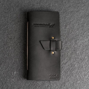 May include: Black leather hunting log with the text "HUNTING LOG" and the year "2024" embossed on the cover. The log features a buckle closure and brass rivets. The log is designed for hunters to record their experiences.