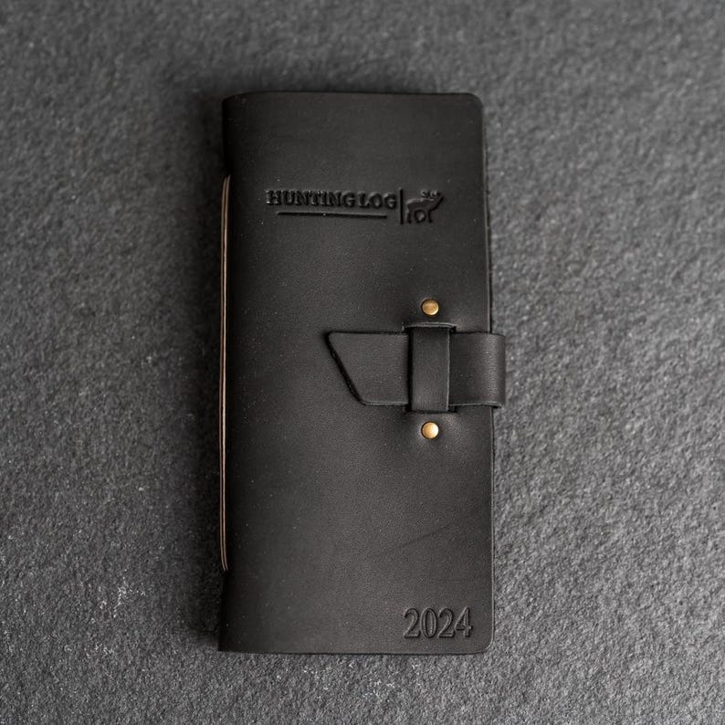 May include: Black leather hunting log with the text "HUNTING LOG" and the year "2024" embossed on the cover. The log features a buckle closure and brass rivets. The log is designed for hunters to record their experiences.