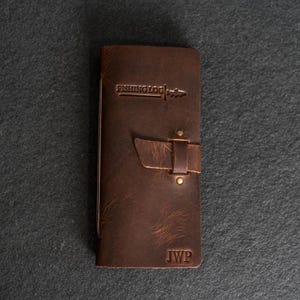 May include: A brown leather fishing log with the text "FISHING LOG" embossed on the front. The log has a strap closure and the initials "JWP" are stamped at the bottom. The leather has a distressed appearance.
