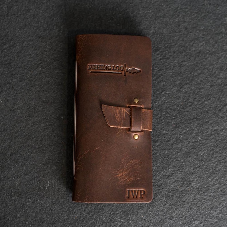 May include: A brown leather fishing log with the text "FISHING LOG" embossed on the front. The log has a strap closure and the initials "JWP" are stamped at the bottom. The leather has a distressed appearance.