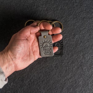 Può includere: Un portachiavi in ​​pelle grigia con le parole "Trust in the Lord" e una grafica di montagna. Il portachiavi è attaccato a un set di anelli portachiavi dorati.