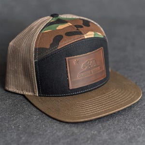 Puede incluir: Gorra de camionero marrón y negra con un estampado de camuflaje en la parte superior y una visera marrón. La gorra presenta un parche de cuero con el emblema de la República de California.