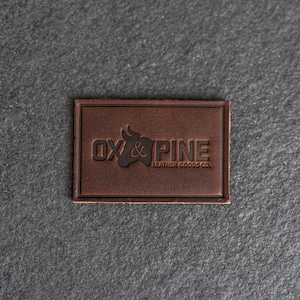 May include: A brown leather patch with the embossed text "OX & PINE LEATHER GOODS CO." The logo features an ox head and pine tree silhouette. The patch has a stitched border and is set against a dark grey background.