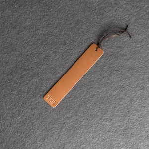 May include: A brown leather bookmark with the initials "TRS" embossed on the bottom. The bookmark has a small hole at the top with a brown string attached. The bookmark is rectangular and lies diagonally on a gray surface.