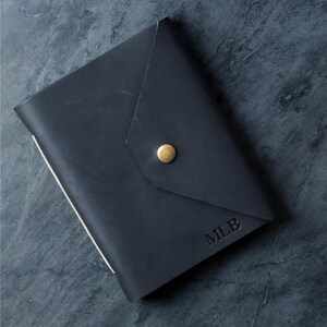 May include: A dark navy blue leather journal cover featuring an envelope-style flap closure secured with a single brass-colored metal stud. The bottom right corner of the cover is embossed with the capital letters MLB in a simple serif font.