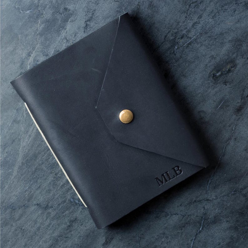 May include: A dark navy blue leather journal cover featuring an envelope-style flap closure secured with a single brass-colored metal stud. The bottom right corner of the cover is embossed with the capital letters MLB in a simple serif font.