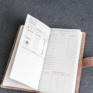 May include: Open leather journal with a detailed hunt record. The journal's pages are filled with sections for weather, wind, and moon phases. The right page has sections for observations and game harvested. The journal is secured with a snap closure.