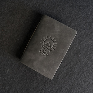May include: A dark gray leather journal with the embossed text "TRUST IN THE LORD" and a mountain and sun design. The journal is rectangular and appears to be closed, with a textured surface. The words "Proverbs 3:5-6" are below the main text.