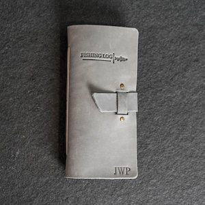 May include: A gray leather fishing log with the text "FISHING LOG" and a fish graphic. The journal has a strap closure with two brass rivets and the initials "JWP" embossed on the bottom. The journal is closed.