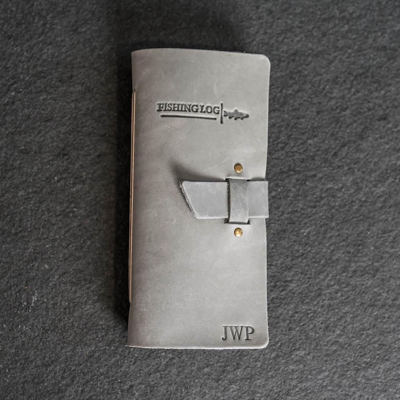 May include: A gray leather fishing log with the text "FISHING LOG" and a fish graphic. The journal has a strap closure with two brass rivets and the initials "JWP" embossed on the bottom. The journal is closed.