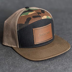Op de afbeelding: Een bruine en zwarte truckerpet met een camouflagepatroon op de bovenkant en een bruine rand. De pet heeft een zwart voorpaneel met een bruin leren patch met de tekst "Anderson". De pet heeft een mesh achterkant.