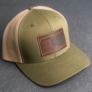 May include: A green and tan trucker hat with a brown leather patch featuring the outline of the state of Oklahoma.