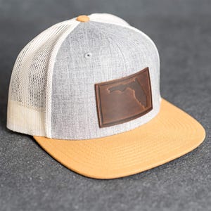 May include: A gray and yellow trucker hat with a leather patch featuring the outline of Florida. The hat has a white mesh back, a yellow brim, and a button on top. The patch is brown.