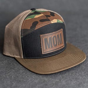May include: A brown and black baseball cap with a camouflage pattern on the top and a mesh back. The cap features a black front panel with a brown leather patch that reads "MOM".