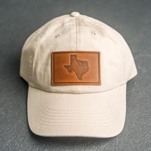 Op de afbeelding: Een beige honkbalpet met een rechthoekige bruine leren patch aan de voorzijde. De patch is voorzien van een reliëf van de omtrek van de staat Texas. De pet heeft een gebogen klep en stikseldetails over de kroon en de klep.