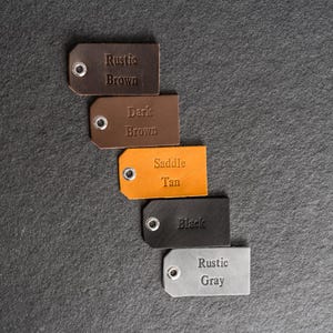 May include: A collection of leather tags in various colors: rustic brown, dark brown, saddle tan, black, and rustic gray. Each tag has a metal eyelet and embossed text indicating the color name. The tags are arranged vertically against a dark background.