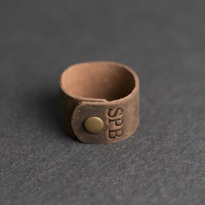 Op de afbeelding: Een bruine leren ring met een gladde, matte afwerking en een ronde, messingkleurige metalen drukknoop. De letters SPB zijn verticaal in het leeroppervlak naast de sluiting gedrukt, wat zorgt voor een eenvoudig en functioneel ontwerp.