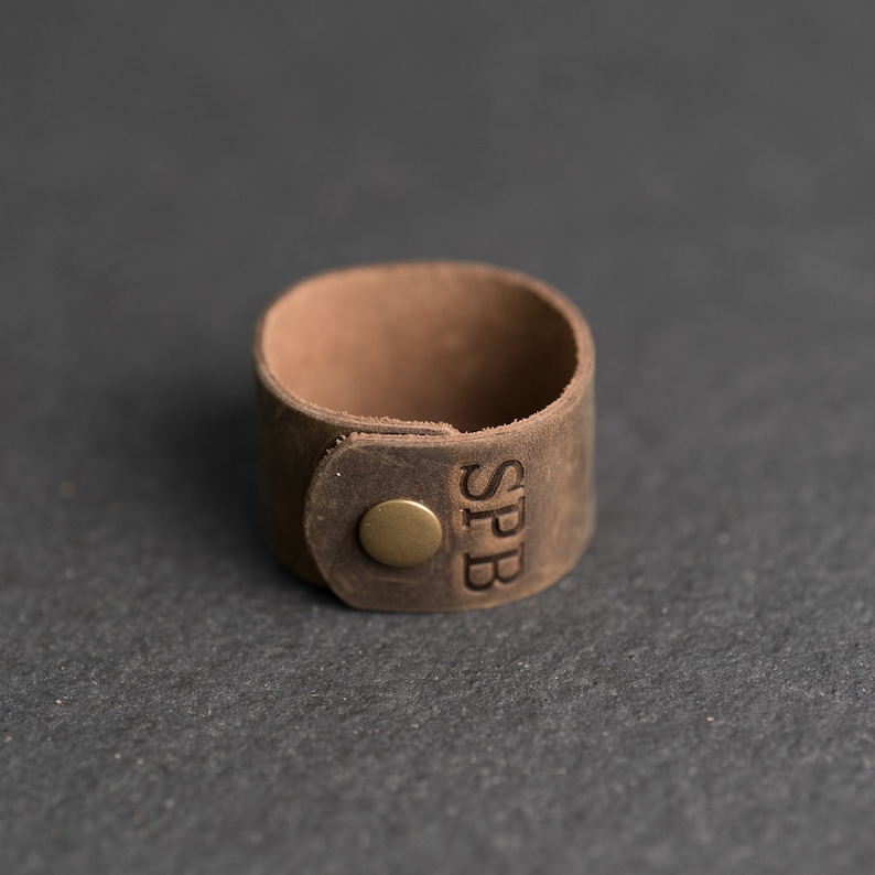 Op de afbeelding: Een bruine leren ring met een gladde, matte afwerking en een ronde, messingkleurige metalen drukknoop. De letters SPB zijn verticaal in het leeroppervlak naast de sluiting gedrukt, wat zorgt voor een eenvoudig en functioneel ontwerp.