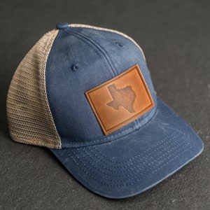 Könnte beinhalten: Eine blaue und braune Trucker-Kappe mit einem Leder-Patch, der den Umriss des Bundesstaates Texas zeigt.