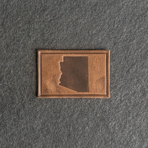 Puede incluir: Un parche de cuero marrón con el contorno del estado de Arizona.