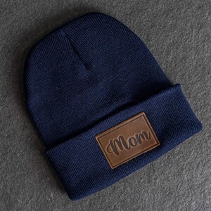 May include: A navy blue knitted beanie hat with a brown leather patch. The patch displays the word "Mom" in a cursive script. The hat is a classic style, suitable for cold weather.