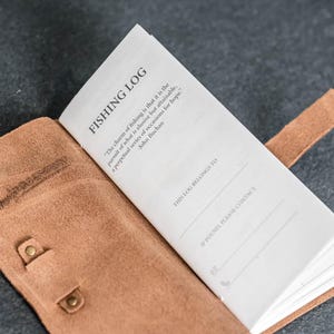 May include: Open leather fishing log book with the title "FISHING LOG" printed at the top. The book is open to a page with a quote by John Buchan. The leather cover is tan with a snap closure.