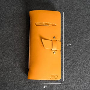 May include: A mustard-colored leather fishing log with the text "FISHING LOG" embossed on the front. The log is 8 inches tall and 4 inches wide, with a buckle closure and the initials "JWP" at the bottom.