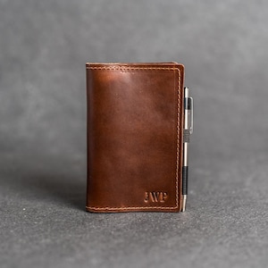 May include: A brown leather wallet with the initials "JWP" embossed on the front. A silver pen is inserted in the side of the wallet. The wallet is a rich, medium brown color and appears to be made of high-quality leather.
