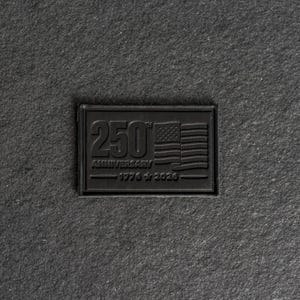 Op de afbeelding: Een zwart rechthoekig embleem met de tekst "250th Anniversary 1776-2026" en een Amerikaans vlagontwerp. Het embleem is in reliëf op een getextureerd, donkergrijs oppervlak.