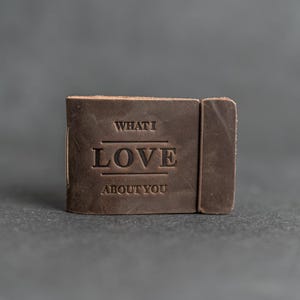 Könnte beinhalten: Ein kleines, rechteckiges, braunes Lederjournal. Auf dem Einband steht der Text "WHAT I LOVE ABOUT YOU". Das Journal ist geschlossen und mit einem Lederband gesichert. Der Hintergrund ist einfarbig grau.