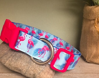 May include: A light blue dog collar with a pink flamingo and palm leaf pattern. The collar has a bright pink plastic buckle and a silver metal D-ring.