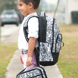 May include: A young person stands on a sidewalk, holding a matching backpack and lunch bag. The bags feature a gray and white digital camouflage pattern. The person is wearing a white shirt, pink shorts, and tan shoes.