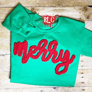 May include: A green sweatshirt with the word "Merry" in red, textured lettering across the front. The sweatshirt has long sleeves and a crew neck. The letters are thick and stand out against the green fabric.