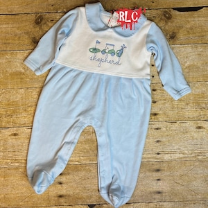 May include: Light blue baby sleeper with long sleeves and footed bottoms. The white bib-style top features embroidered golf-themed graphics and the name "shepherd". The sleeper has a rounded collar and snap closures down the front.