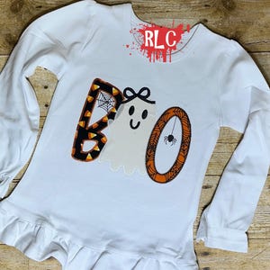 May include: White long-sleeved shirt with a Halloween-themed "BOO" design. The "B" is decorated with candy corn and a spider web, the "O" has an orange spider web, and the second "O" is a ghost with a bow.