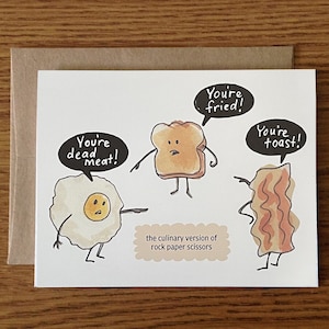 May include: A greeting card with a humorous illustration of a fried egg, a slice of toast, and a piece of bacon. The egg says "You're dead meat!" The toast says "You're fried!" and the bacon says "You're toast!" The text below the illustration says "The culinary version of rock paper scissors."