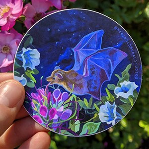 May include: A round sticker featuring a bat with blue wings and a brown body, set against a dark blue starry sky. The bat is surrounded by colorful flowers in shades of purple, white, and green. Text on the sticker reads "@marigoldmuralsstudios".