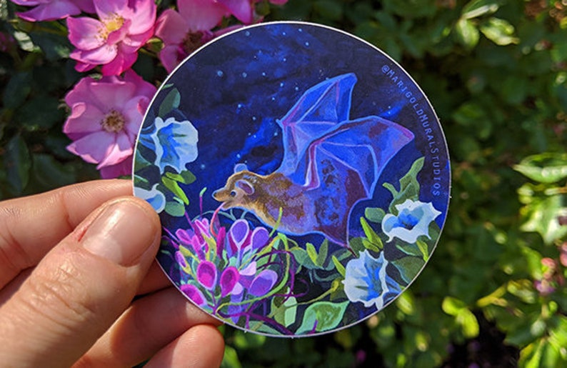 May include: A round sticker featuring a bat with blue wings and a brown body, set against a dark blue starry sky. The bat is surrounded by colorful flowers in shades of purple, white, and green. Text on the sticker reads "@marigoldmuralsstudios".