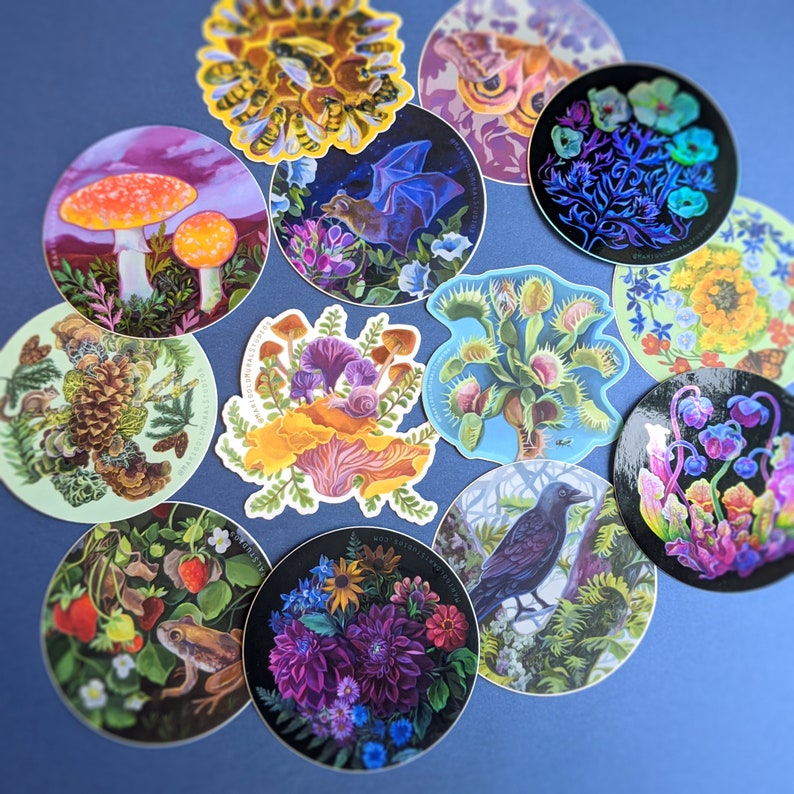 May include: A collection of circular stickers featuring various nature-themed illustrations. Designs include mushrooms, bees, a bat, a Venus flytrap, flowers, and a raven. The stickers have a glossy finish and are arranged on a blue surface.
