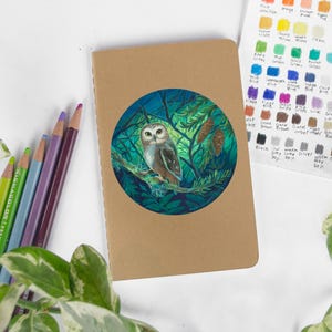 May include: A brown notebook with a circular illustration of an owl perched on a branch within a forest scene. Colored pencils in green, blue, purple, and brown are arranged to the left. A color chart is visible.