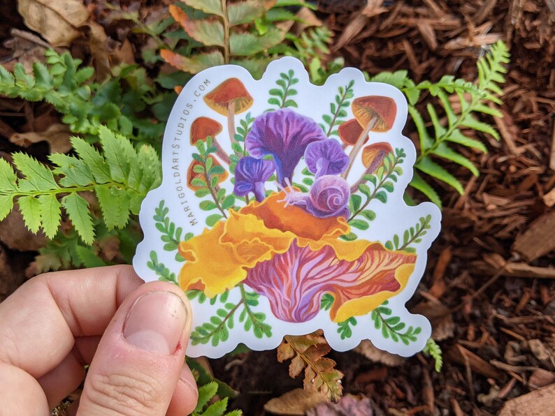 May include: A colorful sticker featuring a whimsical illustration of mushrooms, a snail, and foliage. The mushrooms have brown caps and purple stems, with a snail resting on a large orange and purple mushroom. The sticker has the text "marigoldartstudios.com".