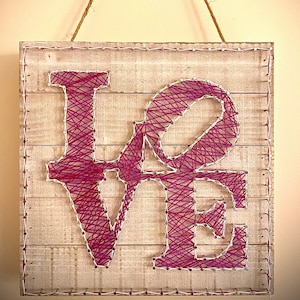 May include: A wooden sign with the word "LOVE" spelled out in pink and white string art. The sign is hanging from a rope and has a rustic, farmhouse style.