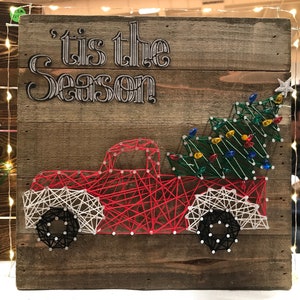 May include: A red truck with a Christmas tree in the back. The truck is made of string art and has white tires. The background is a wooden board with the words "'tis the season" written on it.