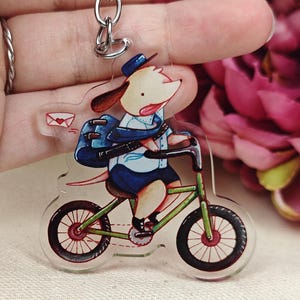 Könnte beinhalten: Ein klarer Acryl-Schlüsselanhänger mit einem Cartoon-Hund, der ein grünes Fahrrad fährt. Der Hund trägt eine blaue Uniform und einen Hut, mit einem Rucksack und einem Brief. Der Schlüsselanhänger hat einen silbernen Ring.