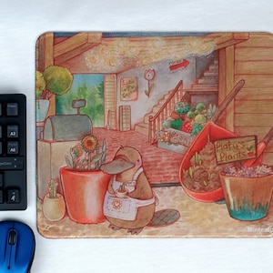 Könnte beinhalten: Mauspad mit einer skurrilen Illustration eines Schnabeltiers in einem Pflanzenladen. Die Szene zeigt Sonnenblumen, eine Schubkarre und ein Schild mit der Aufschrift "Platy's Plants". Das Mauspad hat eine rechteckige Form und eine weiche Stoffoberfläche.